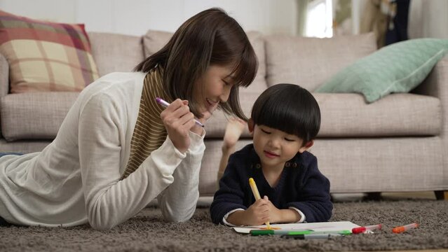 slow motion of loving taiwanese mom lying prone on floor teaching her preschool son to use color pencils drawing on paper at home
