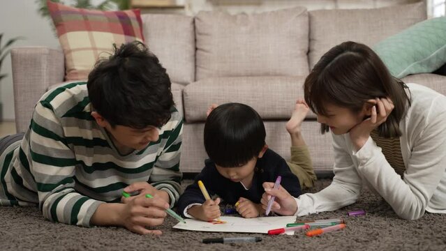 cheerful chinese family of three having fun lying prone on living room floor drawing picture together. the mother laughs at her baby son’s silly talks