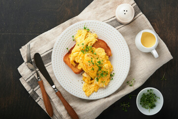 Scrambled Eggs. Fluffy and Buttery scrambled eggs on bread with microgreen radish and hollandaise sauce on white plate over dark old wooden background. Homemade breakfast or brunch meal. Top view.