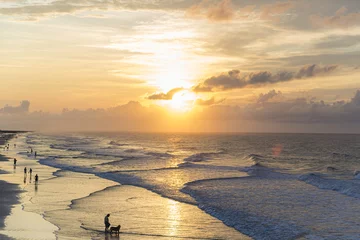 Zelfklevend Fotobehang Prachtig landschap van mensen op het strand met spattende golven onder de bewolkte zonsonderganghemel © Evaldas Sinickas/Wirestock Creators