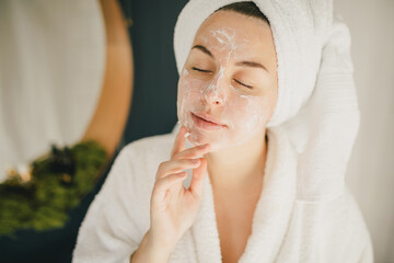 Young beautiful woman wearing bathrobe and towel on her hair applying moistrizing cream on her face. Skin care morning rituals. Beauty routine.