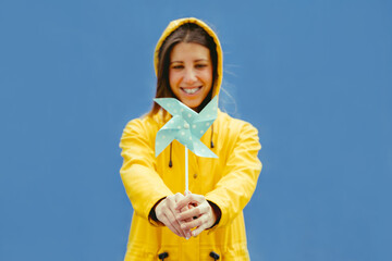 Cropped front view of caucasian woman holding a blue windmill. Horizontal portrait of woman holding funny windmill in yellow raincoat isolated on blue background. Spring conceptual backgrounds.