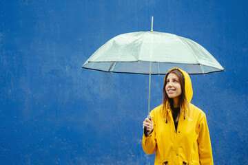 Mid waist portrait of caucasian woman in yellow clothes under rain with umbrella. Horizontal view of caucasian happy woman outdoors holding umbrella on blue background. Rain lifestyle concept