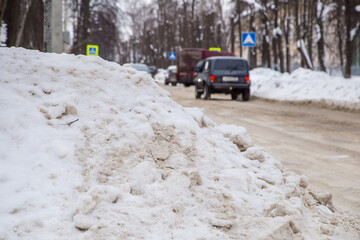 A deep large snowdrift by the road against the backdrop of a city street. On the road lies dirty snow in high heaps. Urban winter landscape. Cloudy winter day, soft light.