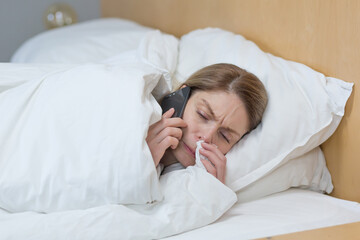 Close-up photo of a sick woman at home, has a bad runny nose and cold, lies in bed covered with a...