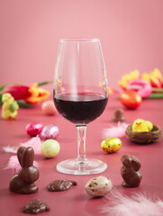 A glass of red wine with Easter chocolate such a rabbit, eggs with festive atmosphere and Easter decorations as a little chick, nest with colorful tulips and pink background 
