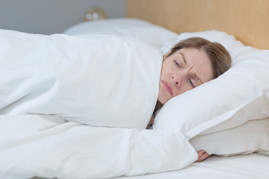 Close-up photo of a sleepless and upset woman lying in bed under a blanket, trying to fall asleep