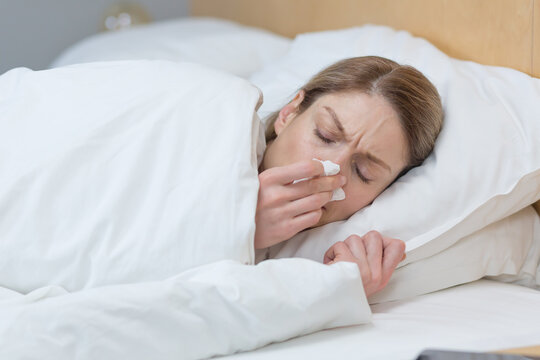 Close-up photo of a sick woman lying under a blanket in bed at home, has a runny nose and cold