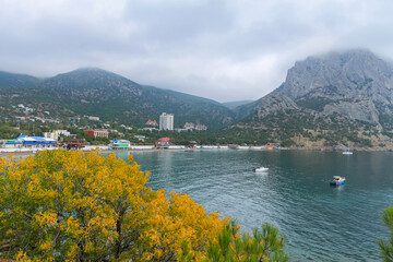Fototapeta premium The sea bay with blue water, boats and yachts. In the foreground yellow tree foliage. In the background of the mountain and cloudy sky. Taken from the Golitsyn Trail. Novy Svet, Crimea