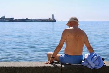 An elderly man in shorts and a cap, without a shirt is sitting on the concrete parapet of the...