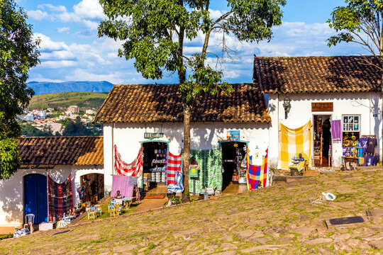 Casebres tradicionais do interior de Minas Gerais com telhados de cer&acirc;mica e artigos coloridos &agrave; venda pendurados na varanda, em um cen&aacute;rio de campo e vegeta&ccedil;&atilde;o