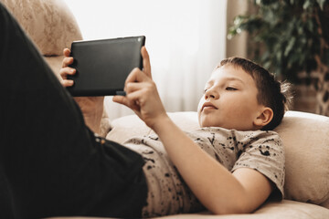 Teenage boy lieing on couch using tablet and playing video games on internet online at cozy home.