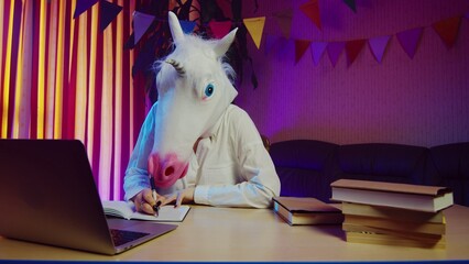 A woman in a unicorn mask sits on a chair at a table and makes notes in her diary. Multicolored lighting. Masquerade © stanis88