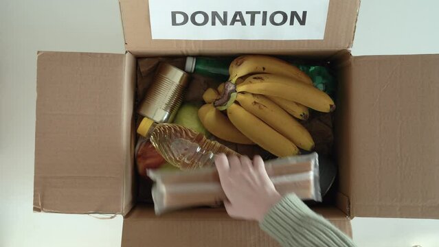 Volunteer putting food In donation box. Delivery woman prepares packing box with food. Donations to refugees in Ukraine. 4k footage. Top view