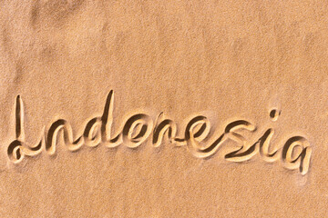 The text "Indonesia" is handwritten on a golden sandy beach near the sea. Next to the text are waves of sand. At the top is copy space. Flat lay photo texture.