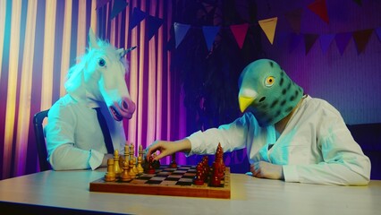A young man and a woman wearing pigeon and unicorn masks are playing chess at a table. Multicolored...