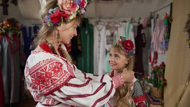 Smiling Ukrainian woman caressing long hair of happy girl in traditional embroidered dress. Beautiful young mother talking with charming daughter indoors in slow motion. Family concept