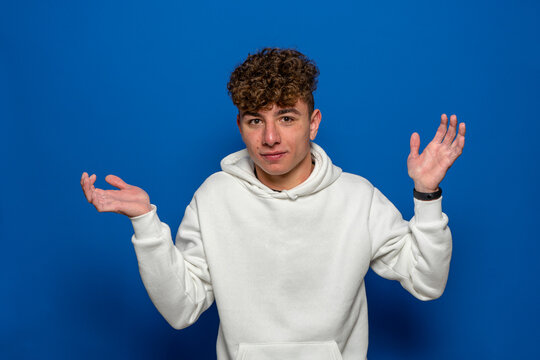 Young handsome brunette man with curly hair wearing white sweatshirt over blue background clueless and confused expression with arms and hands raised. Doubt concept.