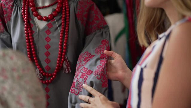 Unrecognizable blurred women caressing embroidered national costume laughing choosing new dress. Two friends standing in Ukrainian craft shop indoors talking smiling