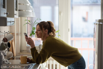 Woman standing in the kitchen and drinking cup of tea while using smartphone