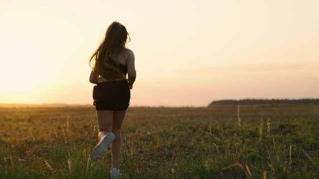 Runner girl runs along green grass. Running after sun. Training jogging. Healthy beautiful girl is engaged in fitness, jogging in country in sun. Free young woman runs in summer park at sunset.