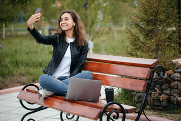 Portrait of a smiling young girl-woman sitting on a bench with a laptop in the park, listening to music with headphones, using a mobile phone for selfies