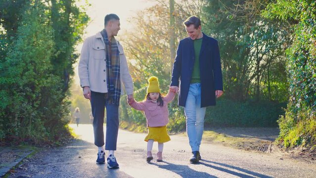 Young daughter holding hands with two fathers as they walk through winter countryside together against flaring sun - shot in slow motion