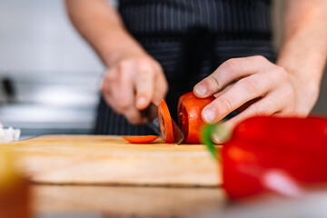 detail of the hands of a young chief man, cutting tomato in a professional kitchen with a knife on a cutting board.