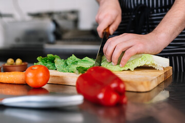 detail of the hands of a young chief man, cutting lettuce in a professional kitchen on a cutting board.