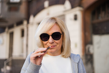 Middle aged blonde European woman smoking heated tabacco e-cigarette outdoors on the street. Alternative smoking way