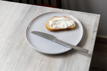 A piece of bread with curd cheese spread on it on a white ceramic plate with a knife on the beige...
