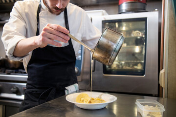 chef manos cocina cpasta fresca tortellini con salsa sirve en plato blanco en interior cocina restaurante con uniforme blanco y mandil negro close-up