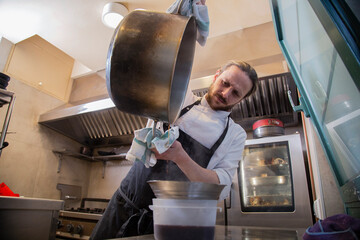 chef rubio con barba en interior de cocina de restaurante filtra salsa de una olla grande metalica...
