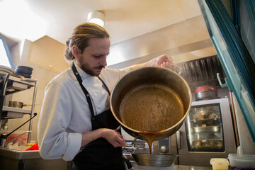 chef rubio con barba en interior de cocina de restaurante filtra salsa de una olla grande metalica...