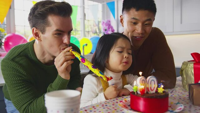 Family with two dads and daughter celebrate parent's 30th birthday with girl blowing party blower at party  - shot in slow motion