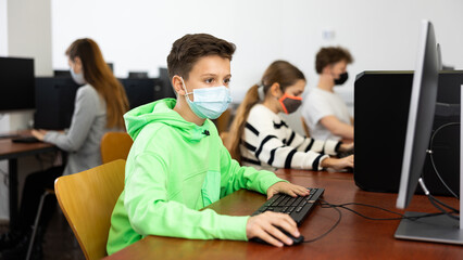 Positive male student wearing face mask working on computer in library. Concept of adult self-education during pandemic