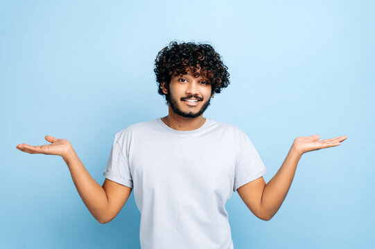 Puzzled Confused Indian Or Arabian Curly-haired Man In Casual Wear, Shrug Shoulders, Looking Questioningly At The Camera, Standing On Isolated Blue Background