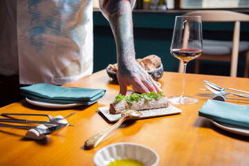 mano de chef sirve pancake de steat tartar con brotes verdes en una mesa de madera en  restaurante con copa de vino tinto y tatuajes close-up