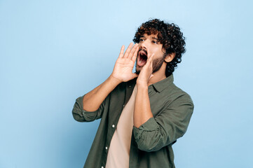 Excited indian or arabic curly haired guy in casual clothes, shouting loudly holding his hands near...