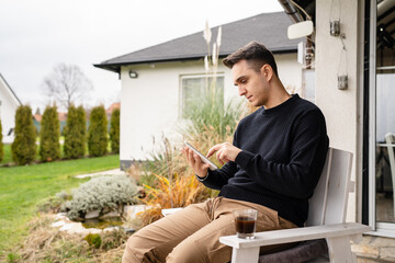 One young adult caucasian man sitting on the chair outdoor in front of his house in day while holding a cup of coffee and digital tablet looking to the side thinking