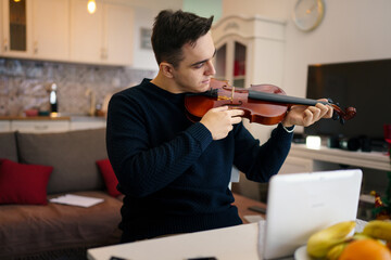 One young adult caucasian man sitting on the chair at home learning to play violin male violinist playing music at his apartment in room alone