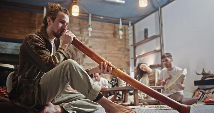 Male hippie playing music with friends. Low angle of man in casual clothes playing didgeridoo while sitting on floor and creating music with friends in loft room