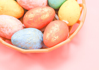 Multi-colored decorative eggs in pastel colors lie in a wicker plastic basket on a light pink.
