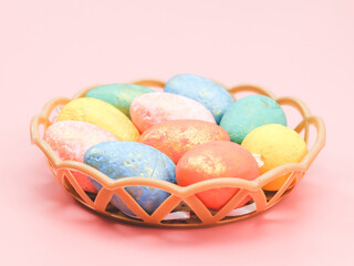 Multi-colored decorative eggs in pastel colors lie in a wicker plastic basket on a light pink.