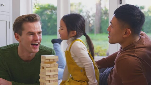 Daughter whispering secret in parent's ear as family with two dads play game stacking wooden bricks into tower - shot in slow motion 