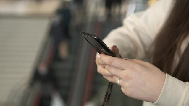 Hands of a young woman using a smartphone in a shopping mall. A young woman is holding a phone in a shopping mall in front of an escalator with people. Close up