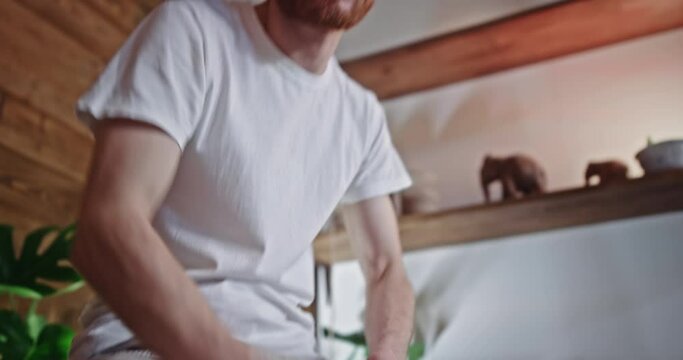 Bearded male hippie playing cajon. Tilt up and tilt down view of glad bearded man in light clothes smiling and playing cajon in daytime at home