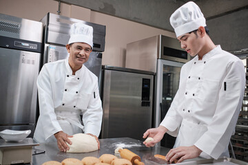 Two professional Asian male chefs in white cook uniforms and aprons are kneading pastry dough and...
