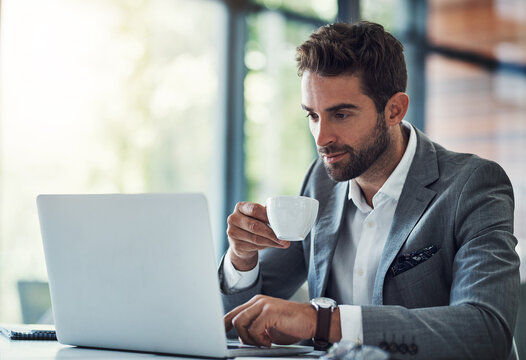 Coffee in one hand, productivity in the other. Shot of a handsome young businessman using a laptop and having coffee at his desk in a modern office.