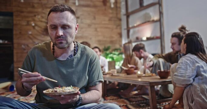 Young man seated on the floor eating Asian food. Young man seated on the floor eating Asian food with chopsticks in gathering of hippie friends indoors in a living room